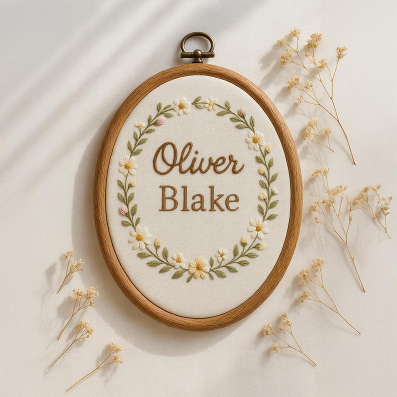 May include: Oval wooden embroidery hoop with the name "Oliver Blake" stitched in brown thread. The name is surrounded by a wreath of white daisies and green leaves. A small brass ring is at the top for hanging.