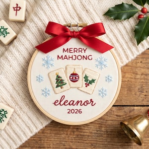 Puede incluir: Un bastidor de bordado redondo de madera con tela blanca que incluye el texto bordado MERRY MAHJONG, eleanor y 2026. El dise&ntilde;o muestra tres fichas de mahjong con un &aacute;rbol de Navidad, una bola y acebo, rodeados de copos azules y un lazo rojo.
