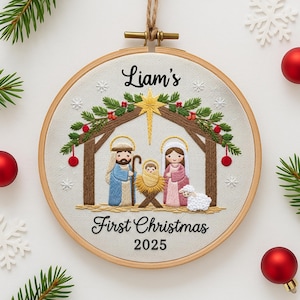 May include: Embroidered Christmas ornament in a wooden hoop. The design features a Nativity scene with the text "Liam's First Christmas 2025". The ornament is decorated with red berries, snowflakes, and a star.