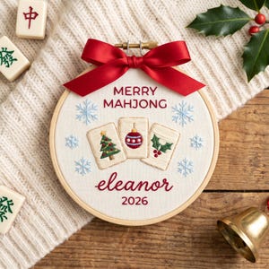 Puede incluir: Un bastidor de bordado redondo de madera con tela blanca que incluye el texto bordado MERRY MAHJONG, eleanor y 2026. El dise&ntilde;o muestra tres fichas de mahjong con un &aacute;rbol de Navidad, una bola y acebo, rodeados de copos azules y un lazo rojo.