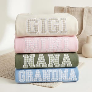 May include: A stack of four crewneck sweatshirts in cream, light pink, olive green, and light blue. Each sweatshirt features large, block-letter text in a gingham pattern that spells out GIGI, MIMI, NANA, and GRANDMA respectively.