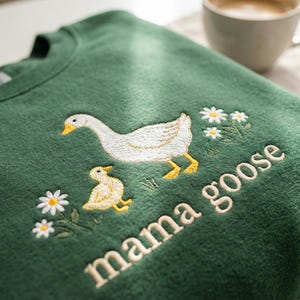 May include: Green sweatshirt with an embroidered design featuring a white goose, a yellow gosling, and daisies. The words "mama goose" are embroidered below the design. A cup of coffee sits in the background.