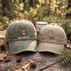 May include: Two olive green baseball caps with embroidered deer designs. One cap says "BIG BUCK" and the other says "LITTLE BUCK". The caps are on a wooden surface with pine needles and pine cones.