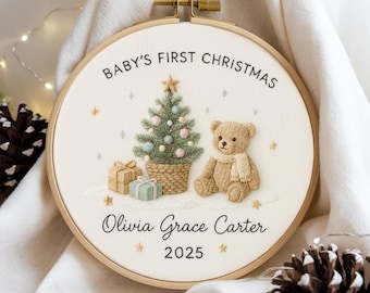 May include: Embroidered hoop art with the text "Baby's First Christmas", a Christmas tree, teddy bear, gifts, and the name "Olivia Grace Carter" with the year 2025. The design is set against a cream-colored fabric, framed by a wooden hoop.