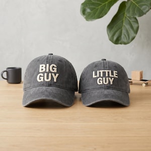 May include: Two gray baseball caps with the phrases "BIG GUY" and "LITTLE GUY" embroidered in white. The caps are displayed on a wooden surface, suggesting a matching set for a parent and child.