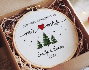 Peut inclure: Un ornement de Noël brodé rond dans un cerceau en bois, présenté dans une boîte en carton avec du papier déchiqueté. L'ornement indique "Our First Christmas as Mr & Mrs" avec un cœur rouge, des sapins et les noms "Emily & Lucas 2024".