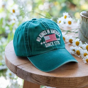 May include: A teal baseball cap with the text "NO KINGS IN AMERICA SINCE 1776" embroidered in white. The design includes an American flag. The cap is sitting on a wooden surface, with daisies and a mug in the background.
