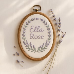 May include: Oval embroidery hoop with the name "Ella Rose" stitched in lavender thread, surrounded by a lavender and green floral wreath. The wooden hoop has a brass-colored clasp. Sprigs of lavender are arranged near the hoop.