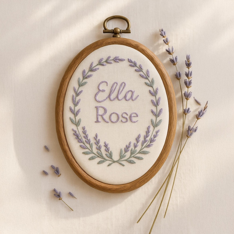 May include: Oval embroidery hoop with the name "Ella Rose" stitched in lavender thread, surrounded by a lavender and green floral wreath. The wooden hoop has a brass-colored clasp. Sprigs of lavender are arranged near the hoop.