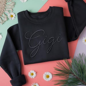 K&ouml;nnte beinhalten: Ein schwarzes Sweatshirt mit dem Namen "Gigi" in einem passenden Ton gestickt. Das Sweatshirt wird auf einer rosa und gr&uuml;nen Oberfl&auml;che pr&auml;sentiert, die mit G&auml;nsebl&uuml;mchen und Gr&uuml;n geschm&uuml;ckt ist. Die &Auml;rmel sind gerollt.