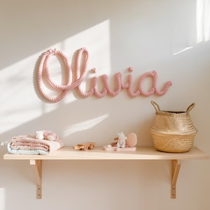 May include: A pink rope name "Olivia" is mounted on a white wall. Below, a wooden shelf holds a folded blanket, toys, and a woven basket. The room is bathed in soft, natural light, creating a cozy atmosphere.