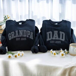 May include: Two navy blue sweatshirts with the words "GRANDPA" and "DAD" in gray block letters, with "EST. 2025" underneath. The sweatshirts are on a white surface with daisies.