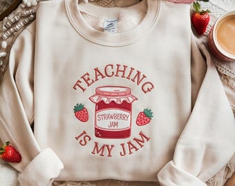May include: Cream-colored sweatshirt with the phrase "TEACHING IS MY JAM" in red script. A graphic of a strawberry jam jar and strawberries is centered on the front. The sweatshirt has long sleeves and a crew neck.