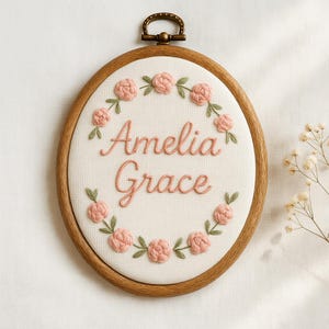 May include: Oval embroidery hoop with the name "Amelia Grace" stitched in pink thread. The name is surrounded by a wreath of pink roses and green leaves. The wooden hoop has a brass-colored metal clasp.