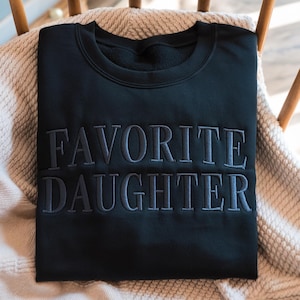 K&ouml;nnte beinhalten: Schwarzes Sweatshirt mit dem gestickten Text "FAVORITE DAUGHTER" in einem dunkleren Grauton. Das Sweatshirt liegt gefaltet auf einer beigen Decke, die &uuml;ber einem Holzstuhl drapiert ist.