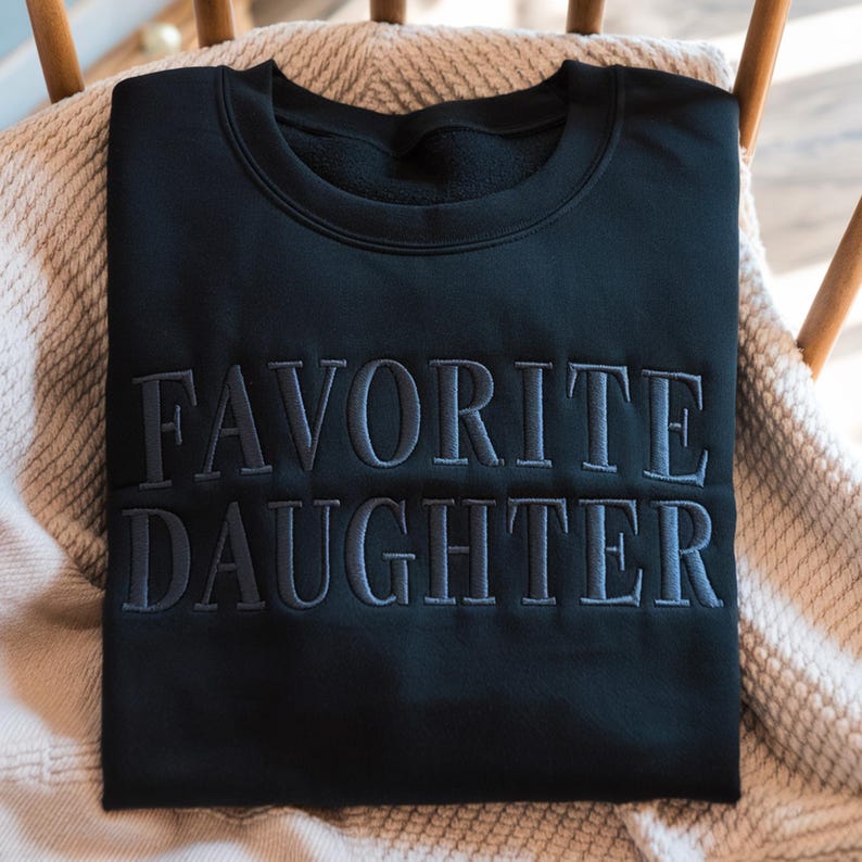 K&ouml;nnte beinhalten: Schwarzes Sweatshirt mit dem gestickten Text "FAVORITE DAUGHTER" in einem dunkleren Grauton. Das Sweatshirt liegt gefaltet auf einer beigen Decke, die &uuml;ber einem Holzstuhl drapiert ist.