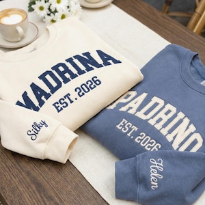 May include: Two crewneck sweatshirts, one cream and one blue, with embroidered text. The cream sweatshirt says "Madrina EST. 2026" in navy blue, with "Silky" on the sleeve. The blue sweatshirt says "Padrino EST. 2026" in white, with "Helen" on the sleeve.
