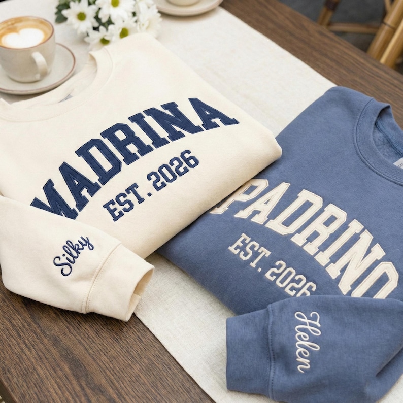 May include: Two crewneck sweatshirts, one cream and one blue, with embroidered text. The cream sweatshirt says "Madrina EST. 2026" in navy blue, with "Silky" on the sleeve. The blue sweatshirt says "Padrino EST. 2026" in white, with "Helen" on the sleeve.