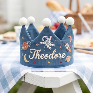 May include: A blue fabric crown with embroidered space-themed designs, including an astronaut, planets, and a rocket. The name "Theodore" is embroidered in cursive. The crown has white pom-poms on top and is sitting on a blue and white checkered surface.