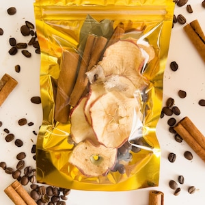May include: A gold zippered pouch filled with dried apple slices, cinnamon sticks, and bay leaves. The pouch is surrounded by coffee beans.