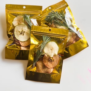 Może przedstawiać: Trzy złote torebki foliowe wypełnione suszonymi plastrami jabłek i gałązkami rozmarynu. Torebki są zamknięte zamkiem błyskawicznym.