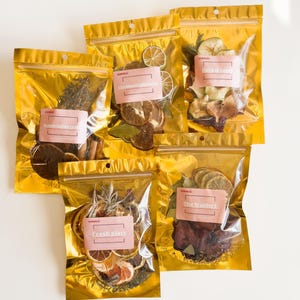 May include: Five gold pouches with different dried fruit and herb blends for tea. The pouches are labeled with the names "Fresh Start", "The Waldorf", "Citrus Mint", "Cranberry Apple", and "Herbal Apple".