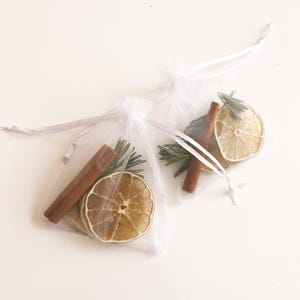 Puede incluir: Dos bolsas de organza blancas llenas de rodajas de cítricos secos, palitos de canela y ramitas de romero. Las bolsas están atadas con una cinta blanca.