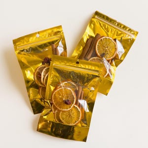 May include: Three gold foil pouches filled with dried orange slices, cinnamon sticks, and other spices. The pouches are sealed with a zipper and have a hanging loop.