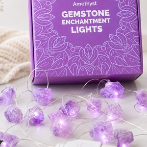 Könnte beinhalten: Eine LED-Lichterkette mit rohen violetten Amethyst-Edelsteinen an einem dünnen silbernen Draht mit weißem USB-Anschluss. Die Lichter liegen vor einer violetten Schachtel mit der Aufschrift Amethyst GEMSTONE ENCHANTMENT LIGHTS.