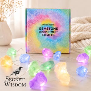 Può includere: Una scatola quadrata con l'etichetta Mixed Rocks GEMSTONE ENCHANTMENT LIGHTS e un motivo mandala arcobaleno. Una catena di luci LED presenta coperture traslucide a forma di roccia in tonalità viola, blu, verde e giallo.