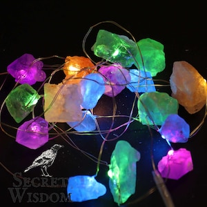 Puede incluir: Una cadena de luces decorativa con cristales de colores. Los cristales son de varios colores, incluyendo morado, verde, naranja y azul. Las luces están ensartadas en un alambre fino, sobre un fondo negro. El texto "Secret Wisdom" está en la esquina inferior izquierda.