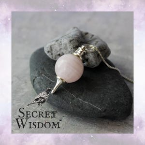 May include: A rose quartz pendulum with a silver chain and tip rests on a dark gray stone. The pendulum is a light pink sphere. The image includes the text "Secret Wisdom" with a raven illustration.