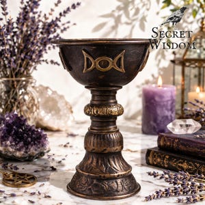 Puede incluir: Un cáliz de color bronce con un cuenco ancho y una base ornamentada. El cuenco presenta un símbolo dorado. El cáliz está rodeado de velas, cristales y lavanda seca, con el texto "Secret Wisdom".