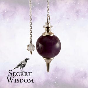 May include: A purple gemstone pendulum with a silver-colored metal cap and chain. A smaller clear crystal pendulum hangs to the left. The image includes a black and white illustration of a raven and the text "Secret Wisdom".