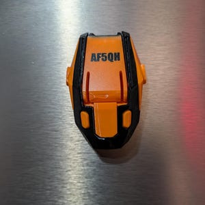 May include: An orange and black device with the text "AF5QH" printed on the top. The device has a geometric design and appears to be made of plastic. The background is a metallic surface.