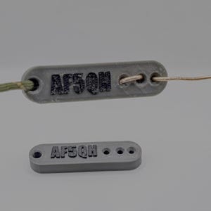 Puede incluir: Se muestran dos etiquetas rectangulares grises con el texto "AF5QH". Una etiqueta está conectada a un cordón verde y marrón y a un alambre fino. La otra etiqueta es más pequeña y tiene tres agujeros.