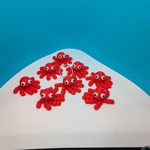 May include: A collection of small, red octopus figurines with white eyes are arranged on a white, curved surface against a bright blue background. The octopus figures have multiple tentacles and are evenly spaced.