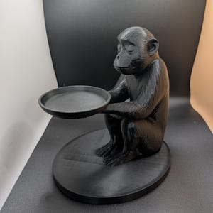 May include: A black monkey figurine holding a round tray. The monkey is seated on a circular base, with its eyes closed. The figurine is made of a matte black material.