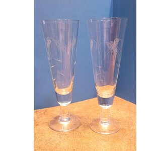 Pode incluir: Duas taças de champanhe transparentes com desenhos de espigas de trigo gravadas. As taças têm uma forma delgada e cónica com uma base redonda e uma haste longa. O design gravado adiciona um elemento decorativo ao vidro.
