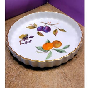 May include: White ceramic pie dish with a scalloped edge and gold trim. The dish is decorated with hand-painted illustrations of plums, oranges, and grapes. The fruit illustrations are in shades of purple, orange, and green.