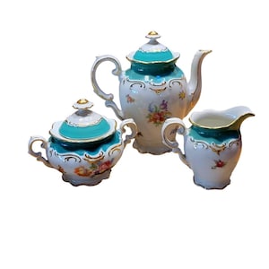 May include: A vintage porcelain tea set comprising a teapot, sugar bowl, and creamer. The set has a white base with teal accents and floral designs. Gold trim adorns the edges. A classic tea set for serving tea.