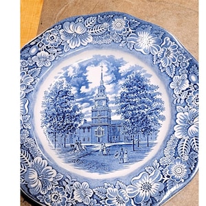 May include: Blue and white decorative plate featuring a historical scene of a building with a clock tower, trees, and figures. The plate has a floral border design. The plate is a square shape with rounded edges.