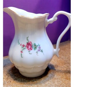 Pode incluir: Jarro de cerâmica branca com borda ondulada e alça. O jarro apresenta um design floral com rosas rosa e vermelhas, folhas verdes e detalhes roxos. A borda é adornada com ouro.