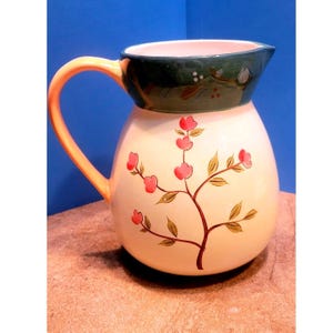 May include: Cream-colored ceramic pitcher with a floral design. The pitcher has a dark green rim and a peach-colored handle. Pink flowers and brown stems decorate the body of the pitcher.