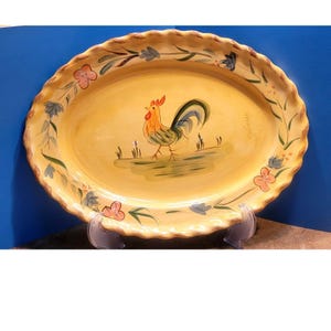 May include: Oval ceramic serving platter with a yellow background and a hand-painted rooster design. The rim is scalloped and decorated with floral and leaf motifs in shades of pink, blue, and green. The platter is supported by a clear plastic stand.