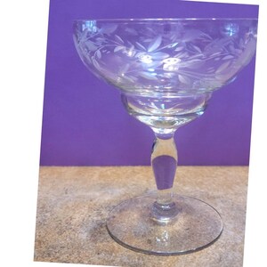 Pode incluir: Um copo de cocktail transparente com um desenho floral gravado em volta da taça. O copo tem uma taça larga e rasa, uma haste curta e uma base circular. A haste tem um detalhe roxo. O fundo é roxo liso.