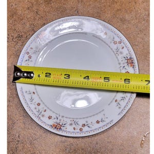 May include: White ceramic plate with a floral pattern and a silver rim. The plate is measured with a tape measure, showing a diameter of approximately 15 cm.