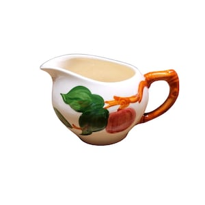 May include: White ceramic creamer with a curved spout and a brown handle. The creamer is decorated with green leaves and red and orange fruit. The handle has a brown, wood-like appearance.