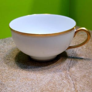 Puede incluir: Una taza de té blanca con borde y asa dorados. La taza tiene una abertura ancha y una base redondeada. El borde dorado añade un toque de elegancia al diseño clásico. La taza de té es probablemente de porcelana o un material similar.