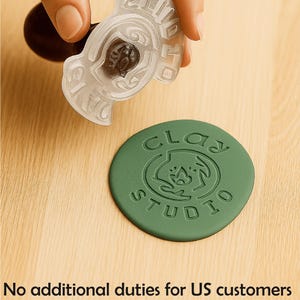 Puede incluir: Un sello decorativo transparente con un mango marrón oscuro se sostiene sobre un sello circular verde. El sello transparente tiene el texto "2017" y "CLAY STUDIO" en relieve. El sello verde también tiene las palabras "CLAY STUDIO" y un logotipo.